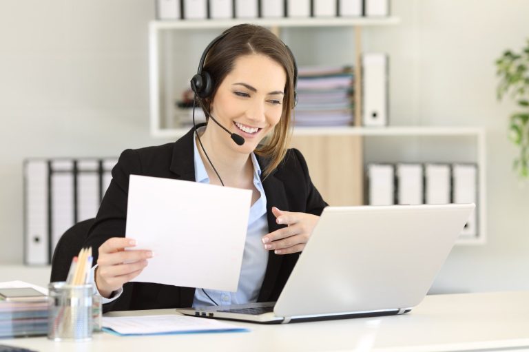 Frau mit Headset, die vor einem Laptop sitzt und ein Dokument in der Hand hält.