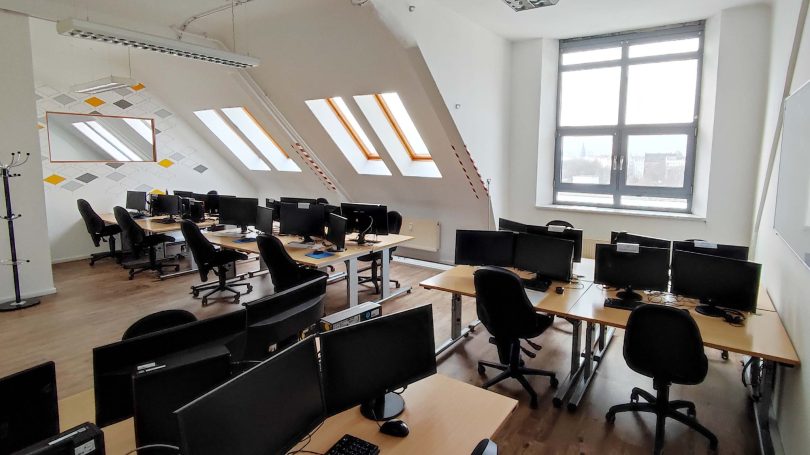 Büro mit mehreren Schreibtischen, Stühlen und Computerarbeitsplätzen in einem lichtdurchfluteten Raum.
