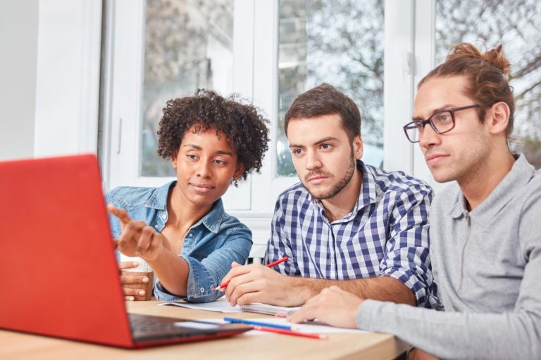 Drei Personen am Tisch schauen auf einen Laptop und diskutieren.