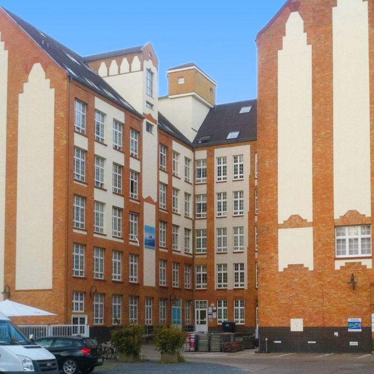 Zwei rote Ziegelgebäude mit spitzen Fassaden und einem Hof zwischen ihnen.